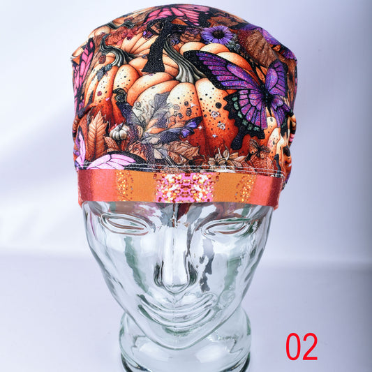 A multicolor autumn-themed fabric head covering or cap with a pumpkin, butterfly, and leaf pattern, displayed on a clear glass mannequin head. The cap features a prominent orange band with a pink and gold patterned center.