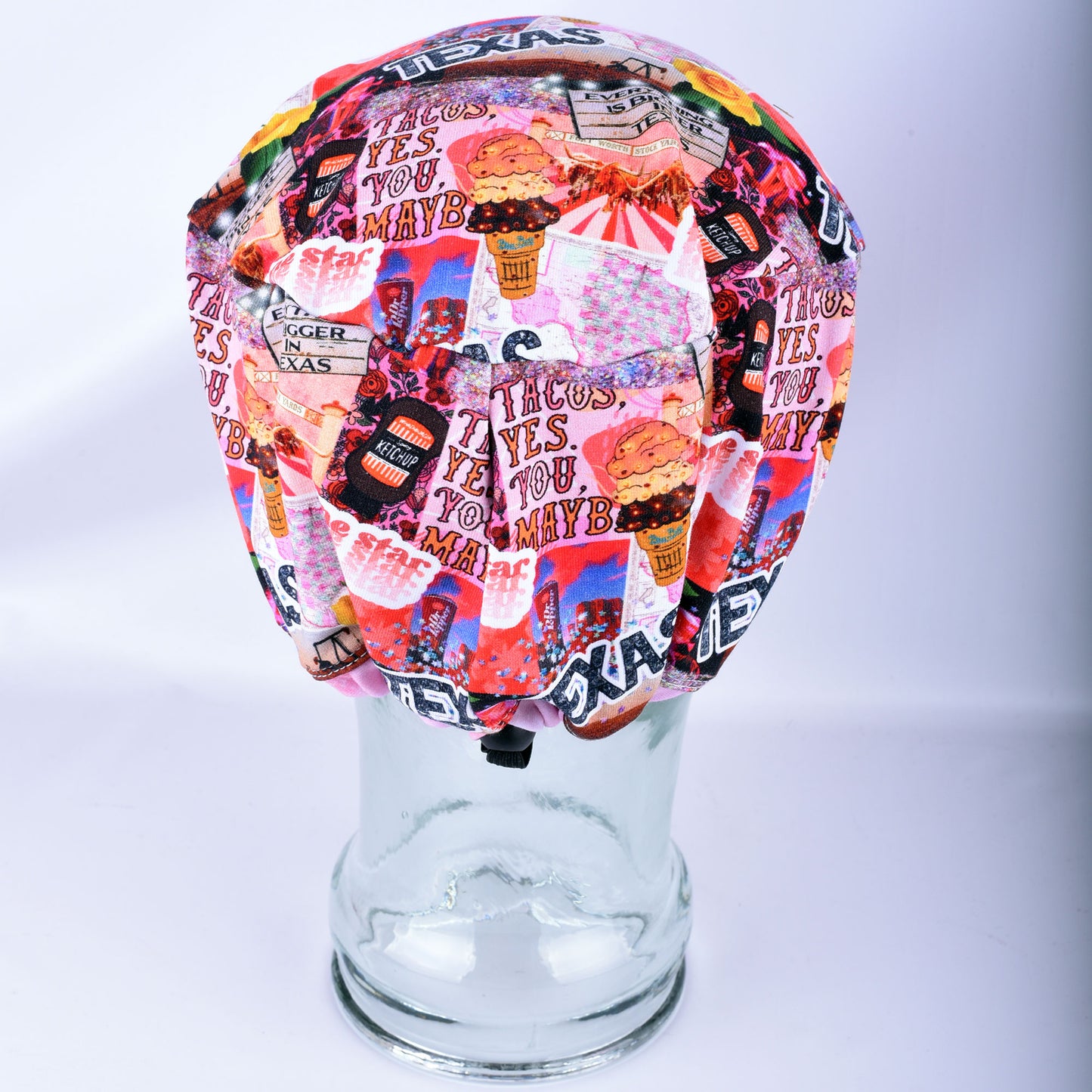 A multicolor scrub cap with a busy collage print featuring 'Texas Yes You Maybe' text, ice cream cones, and retro graphics, displayed on a clear glass mannequin head against a white background.