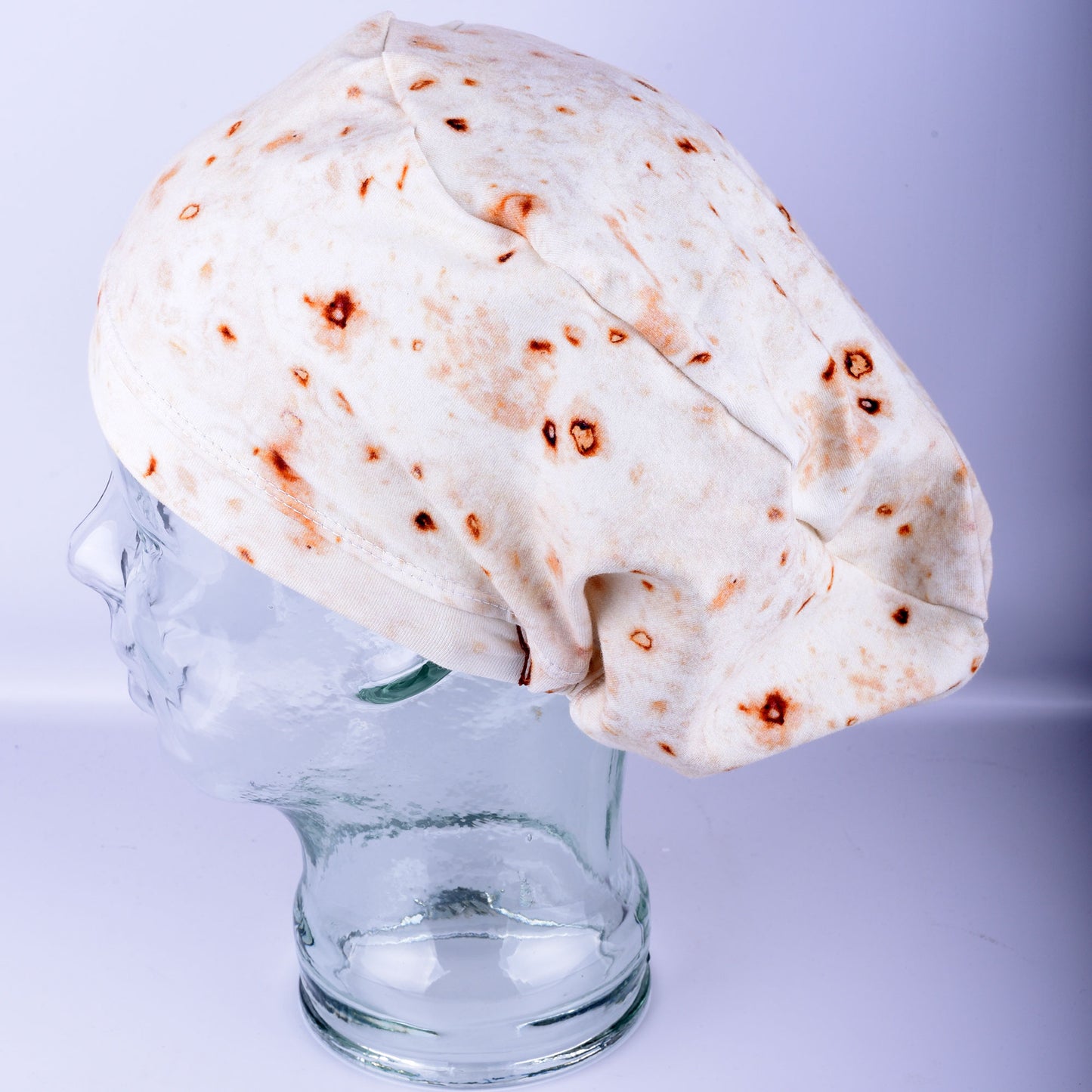 A full coverage scrub cap featuring a realistic flour tortilla print with toasted brown spots, displayed on a white mannequin head.