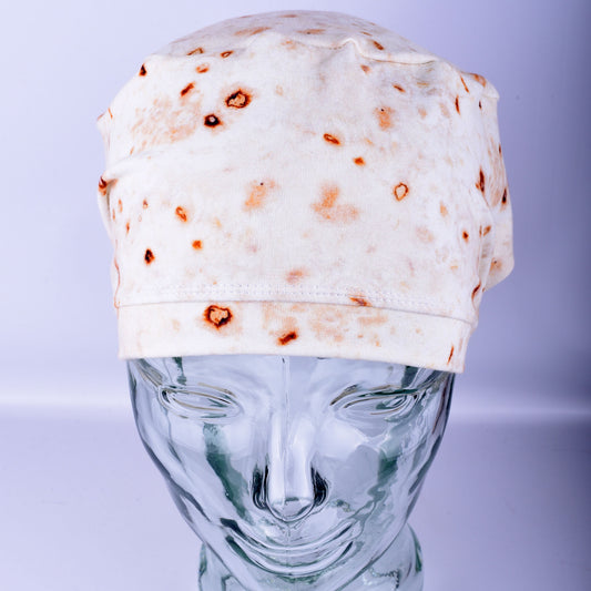 A full coverage scrub cap featuring a realistic flour tortilla print with toasted brown spots, displayed on a white mannequin head.