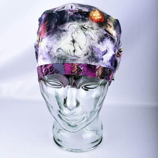 A Halloween-themed surgical scrub cap displayed on a clear glass mannequin head. The cap features a dark, spooky print with a white ghost, a jack-o'-lantern, and purple glittery accents.