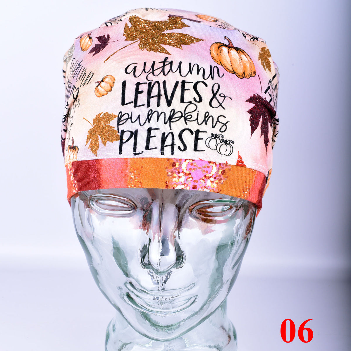 A scrub cap displayed on a clear mannequin head.  The scrub hat has an autumn theme with illustrations of pumpkins, leaves, and the text 'autumn LEAVES & pumpkins PLEASE' on a pink background, with a glittery orange band around the middle.