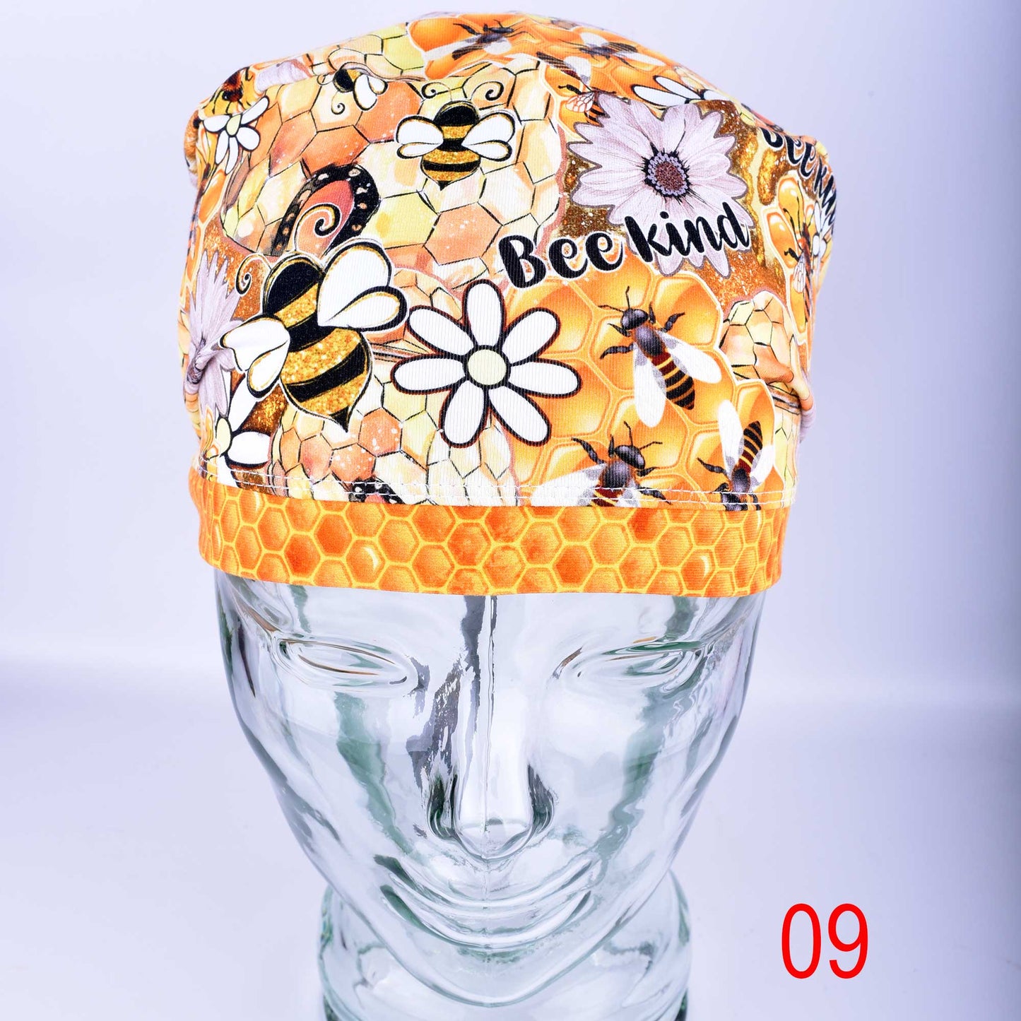 A full-coverage cotton knit medical scrub cap featuring a vibrant "Bee Kind" print with honeycomb patterns, sunflowers, and honeybees on a clean white background. Shown displayed on a clear glass mannequin head.
