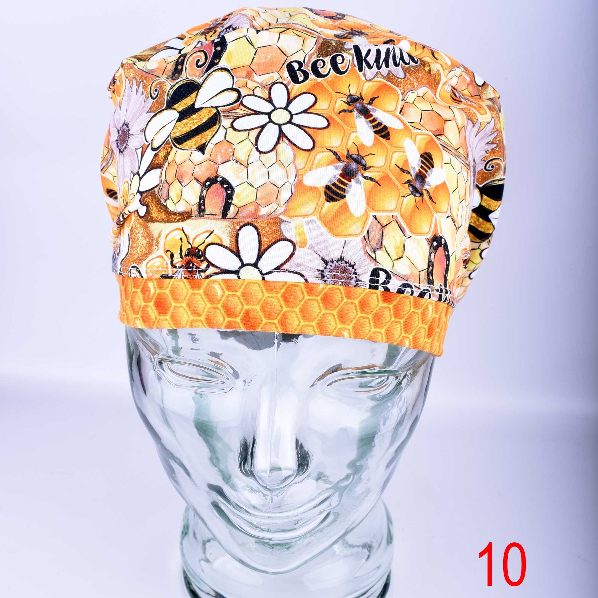 A full-coverage cotton knit medical scrub cap featuring a vibrant "Bee Kind" print with honeycomb patterns, sunflowers, and honeybees on a clean white background. Shown displayed on a clear glass mannequin head.