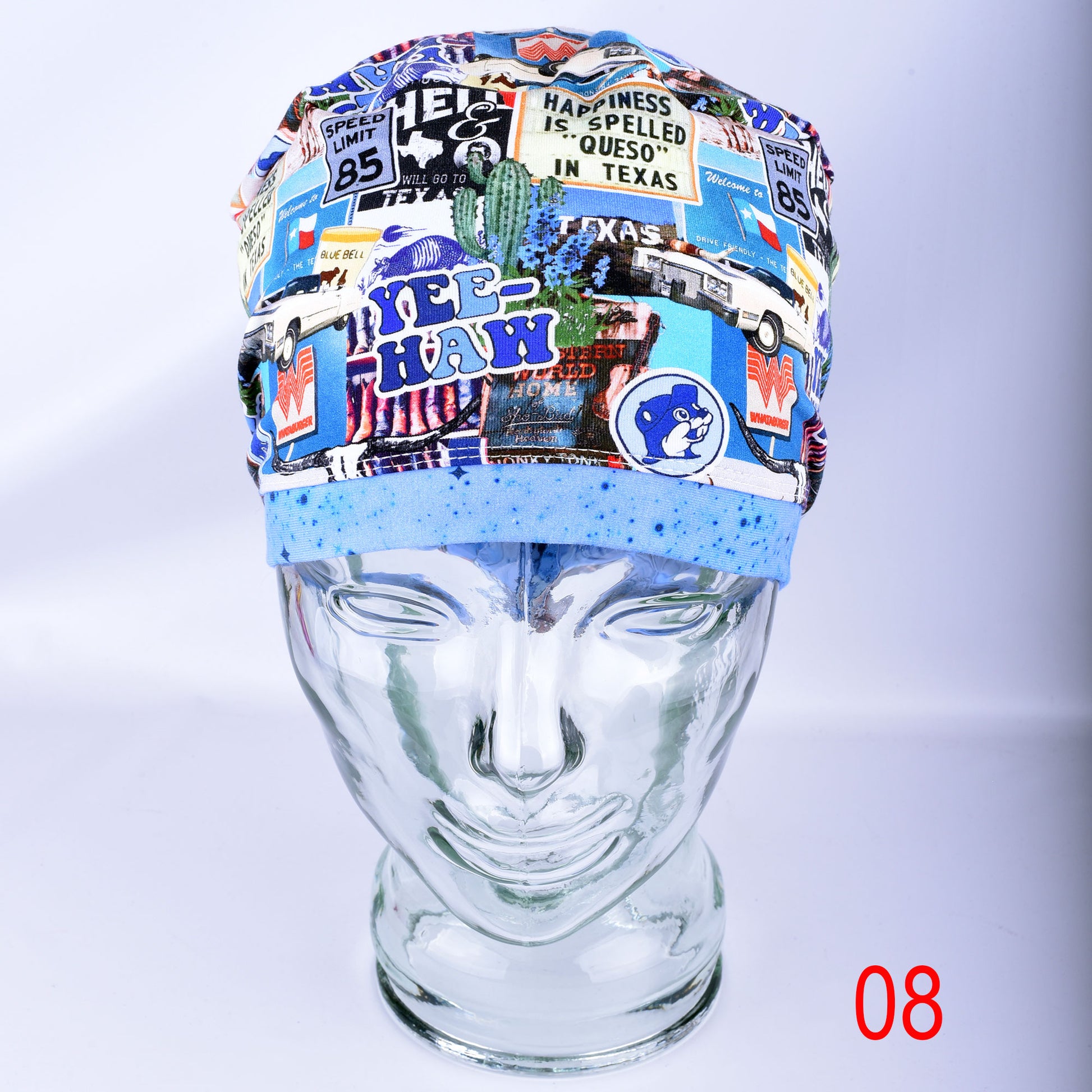 A multicolor novelty scrub cap with a collage of vintage-style travel signs, stickers, and text including 'HAPPINESS IS SPELLED QUESO IN TEXAS' and 'YEE-HAW', displayed on a clear glass mannequin head against a light gray background.