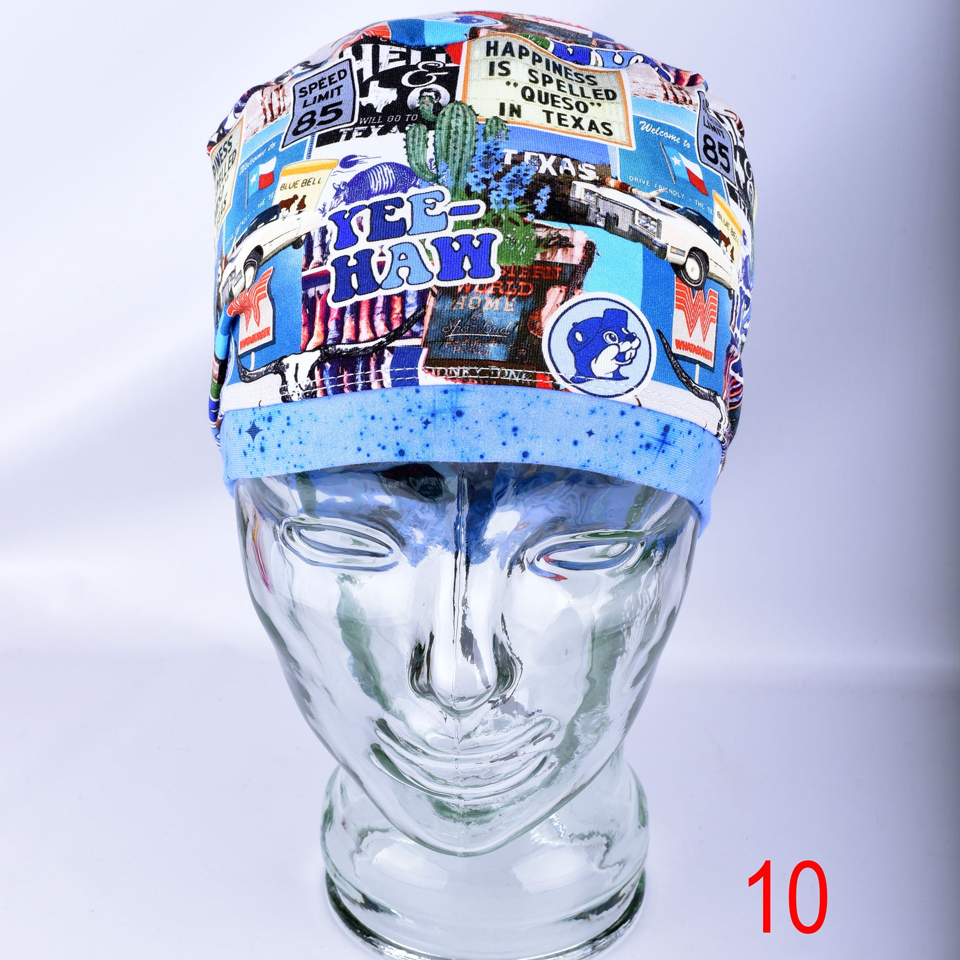 A multicolor collage-print surgical scrub cap displayed on a clear glass mannequin head. The cap features a blue brim with blue glitter and a busy background collage of medical-themed graphics, including a Texas state outline, a cactus, and road signs.