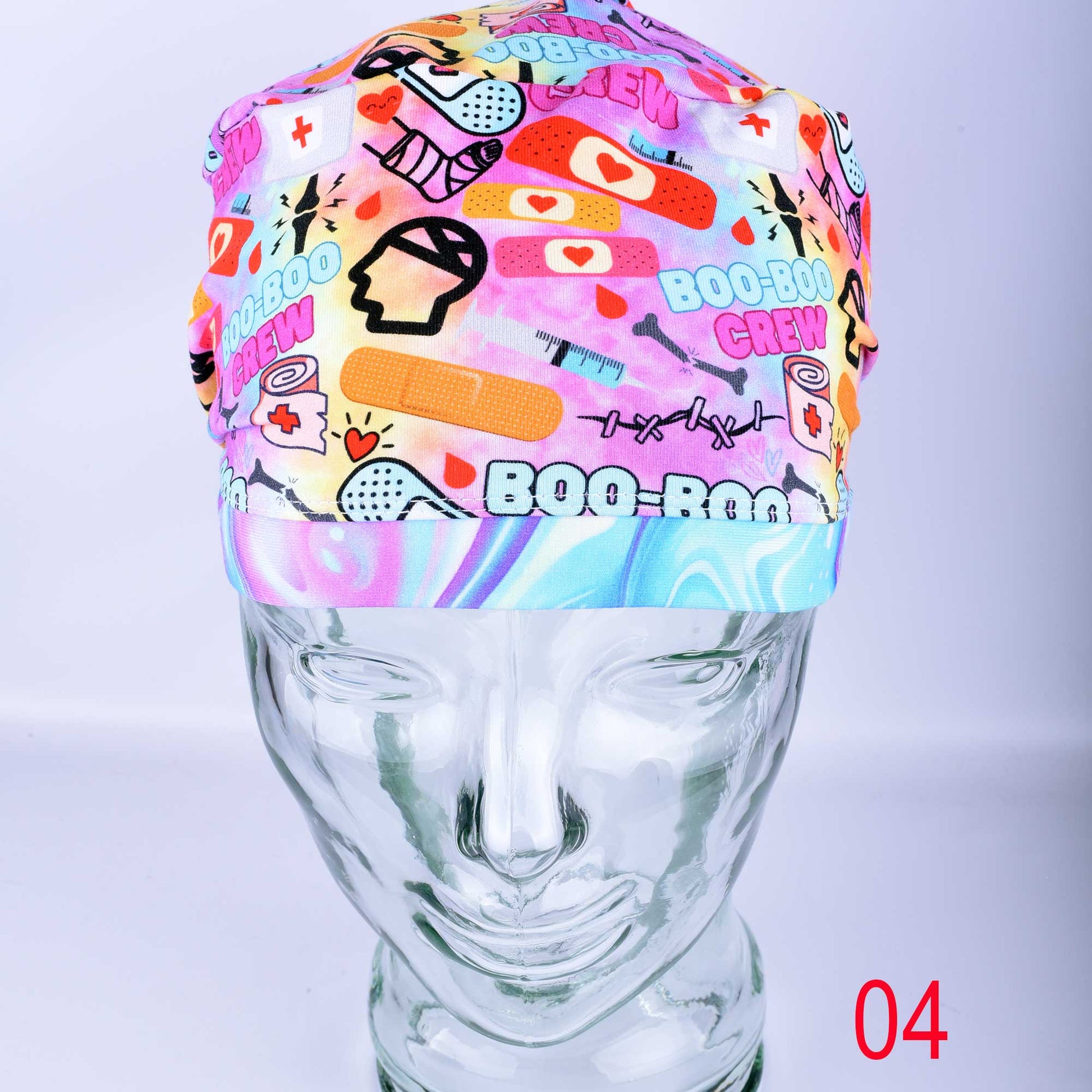 A colorful Boo Boo Crew full coverage scrub cap displayed on a clear mannequin head against a light gray background. The medical hat features a vibrant pink, purple, and yellow background pattern with playful medical graphics, including cartoon bandages, a broken leg cast, a skull, and "Boo-Boo Crew" text, finished with a matching swirled tie-dye style brim.