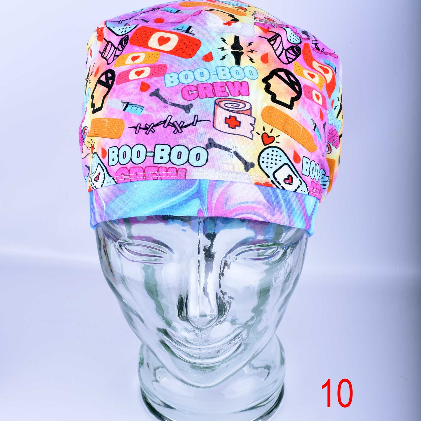 A colorful Boo Boo Crew full coverage scrub cap displayed on a clear mannequin head against a light gray background. The medical hat features a vibrant pink, purple, and yellow background pattern with playful medical graphics, including cartoon bandages, a broken leg cast, a skull, and "Boo-Boo Crew" text, finished with a matching swirled tie-dye style brim.