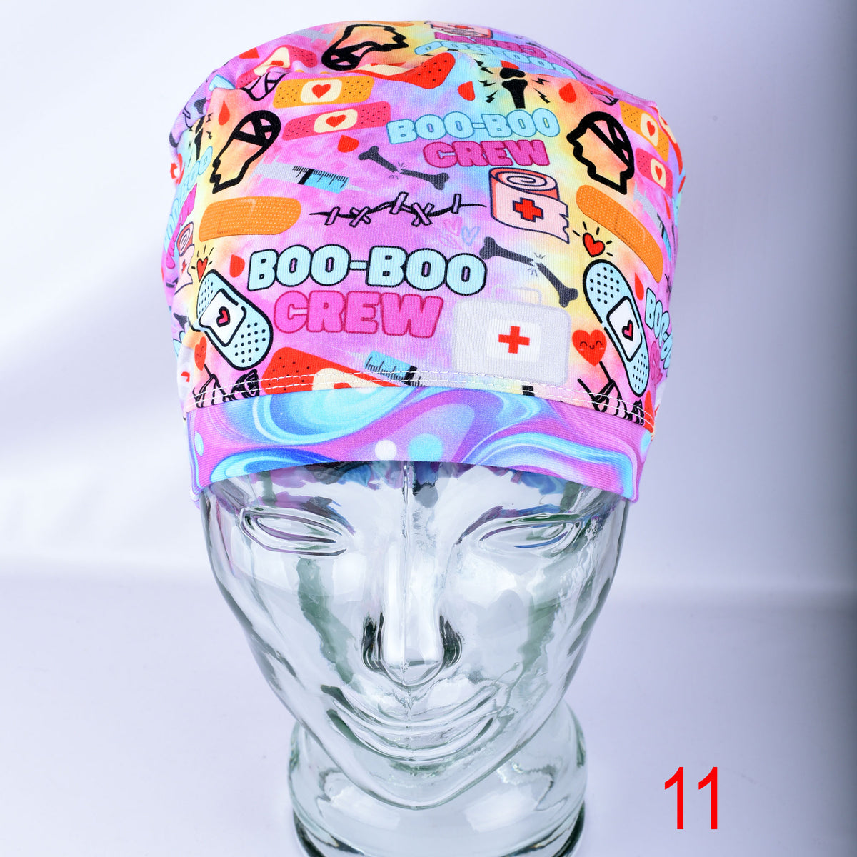 A multicolor scrub cap with a 'BOO-BOO CREW' medical-themed print, displayed on a clear glass mannequin head against a light gray background.