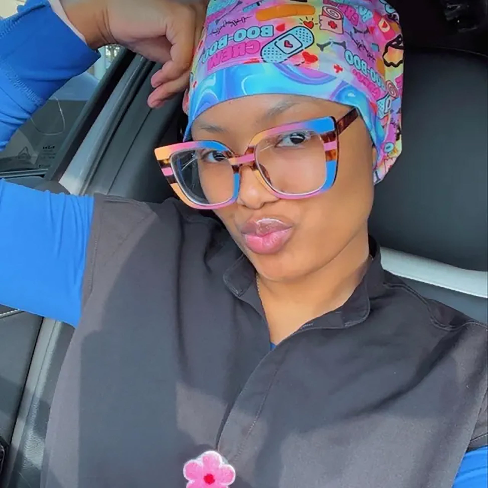 Customer wearing the colorful Boo Boo Crew scrub cap featuring pastel bandages and medical graphics, styled with pink and blue cat-eye glasses and dark gray scrubs.