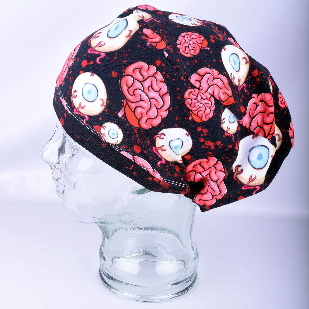 Side profile of a black full-coverage surgical scrub cap featuring a spooky brain and eyeball graphic print on a glass mannequin head.