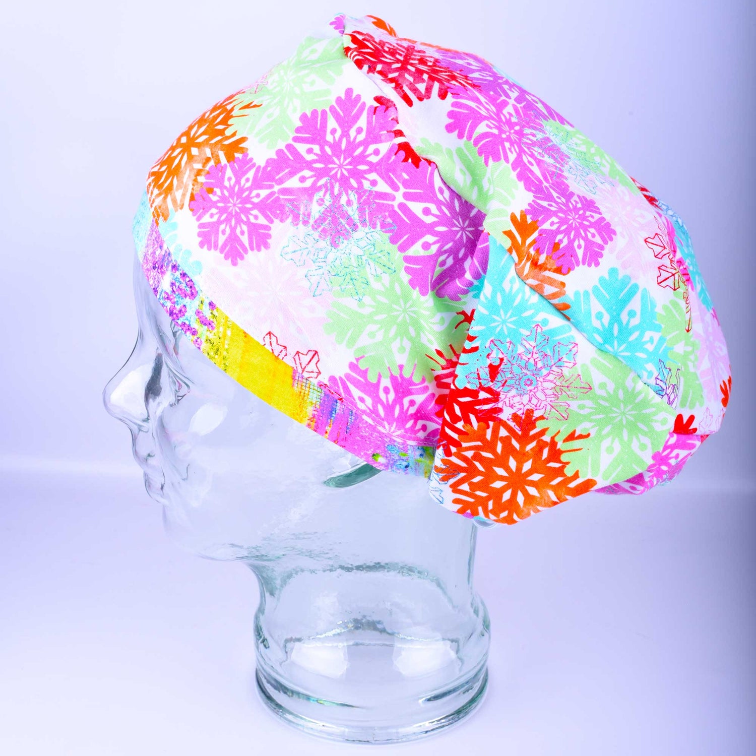 A festive Colorful Snowflakes full coverage scrub cap displayed on a clear mannequin head against a light blue-gray background. The medical hat features a vibrant pink fabric densely patterned with intricate, multi-colored geometric snowflakes in shades of neon green, bright orange, teal, and purple, beautifully finished with a bright yellow brim.