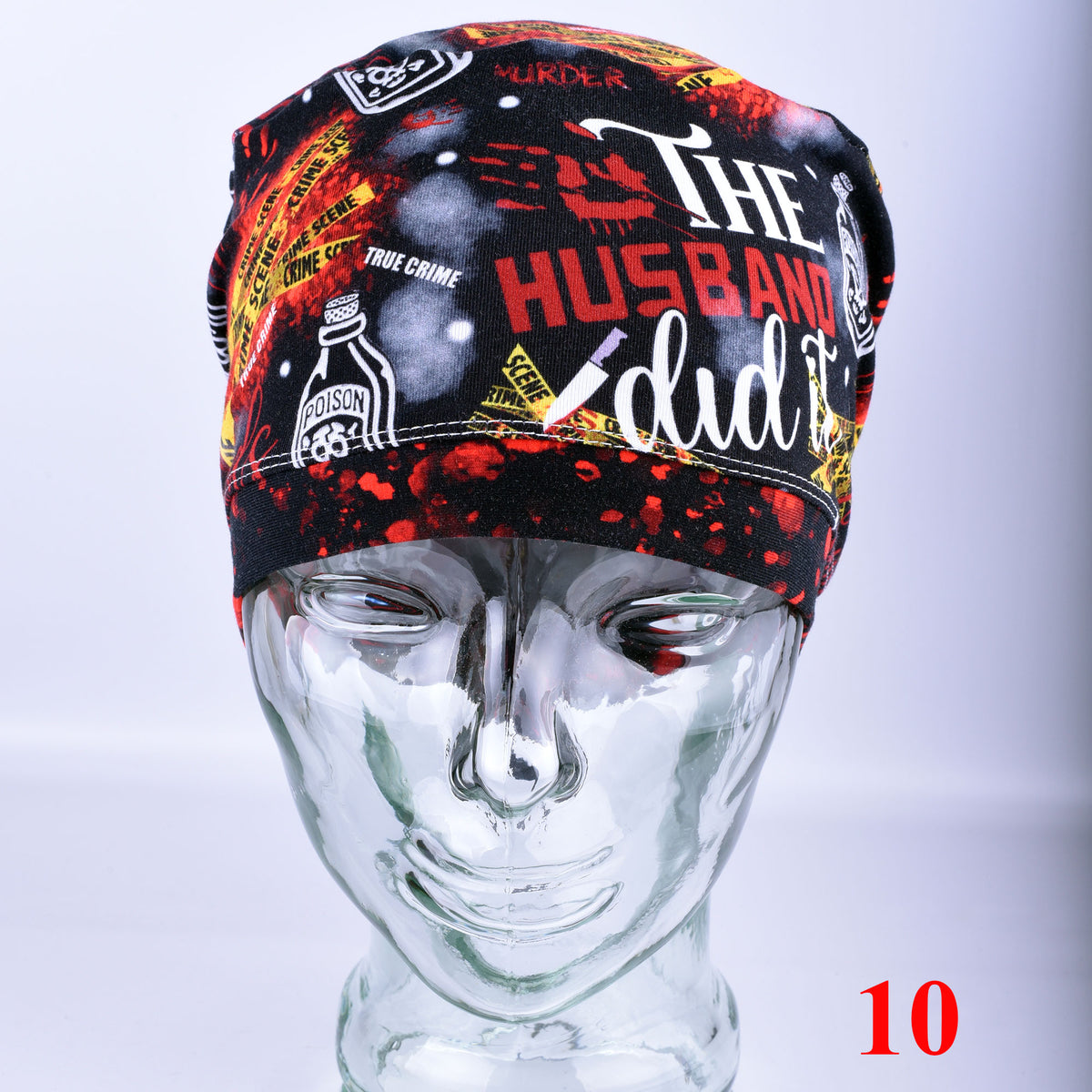 A black scrub cap with a bold graphic print featuring the words 'THE HUSBAND did it' in white and red, along with crime-themed illustrations, displayed on a clear mannequin head against a light gray background.