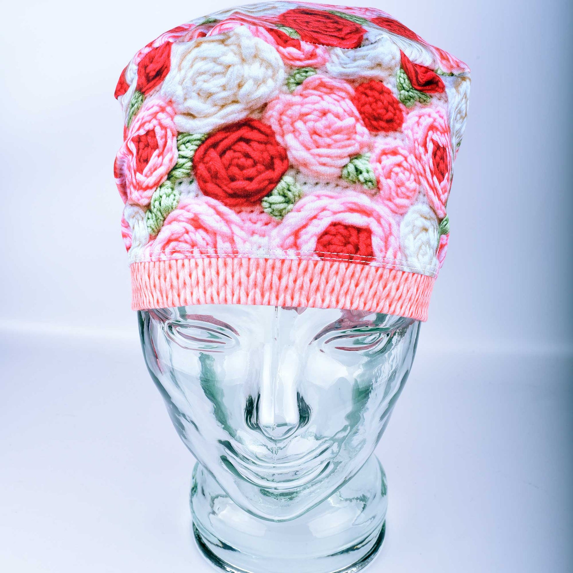 A full-coverage Euro-style scrub cap featuring a vibrant "Crochet Roses" pattern with pink and red floral motifs on a white background, displayed on a glass headform.
