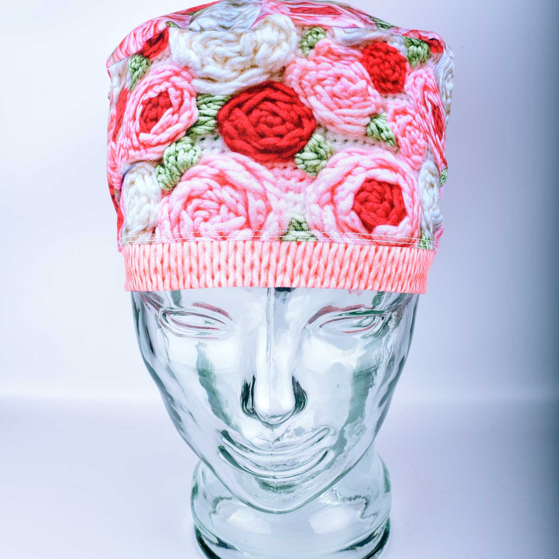 A full-coverage Euro-style scrub cap featuring a vibrant "Crochet Roses" pattern with pink and red floral motifs on a white background, displayed on a glass headform.
