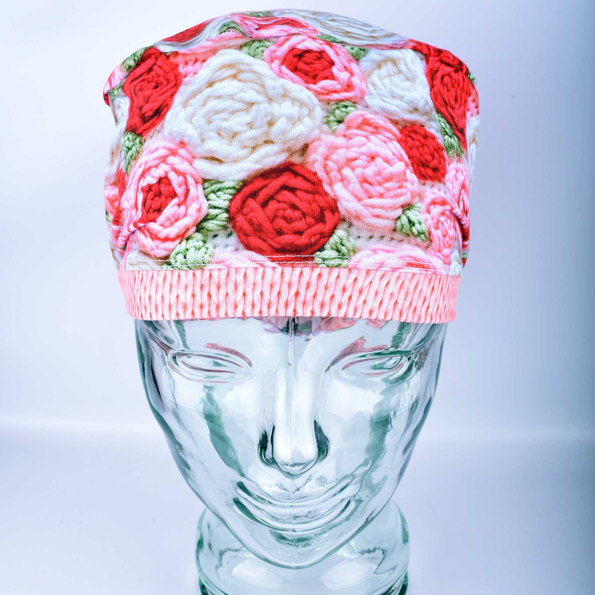 A full-coverage Euro-style scrub cap featuring a vibrant "Crochet Roses" pattern with pink and red floral motifs on a white background, displayed on a glass headform.