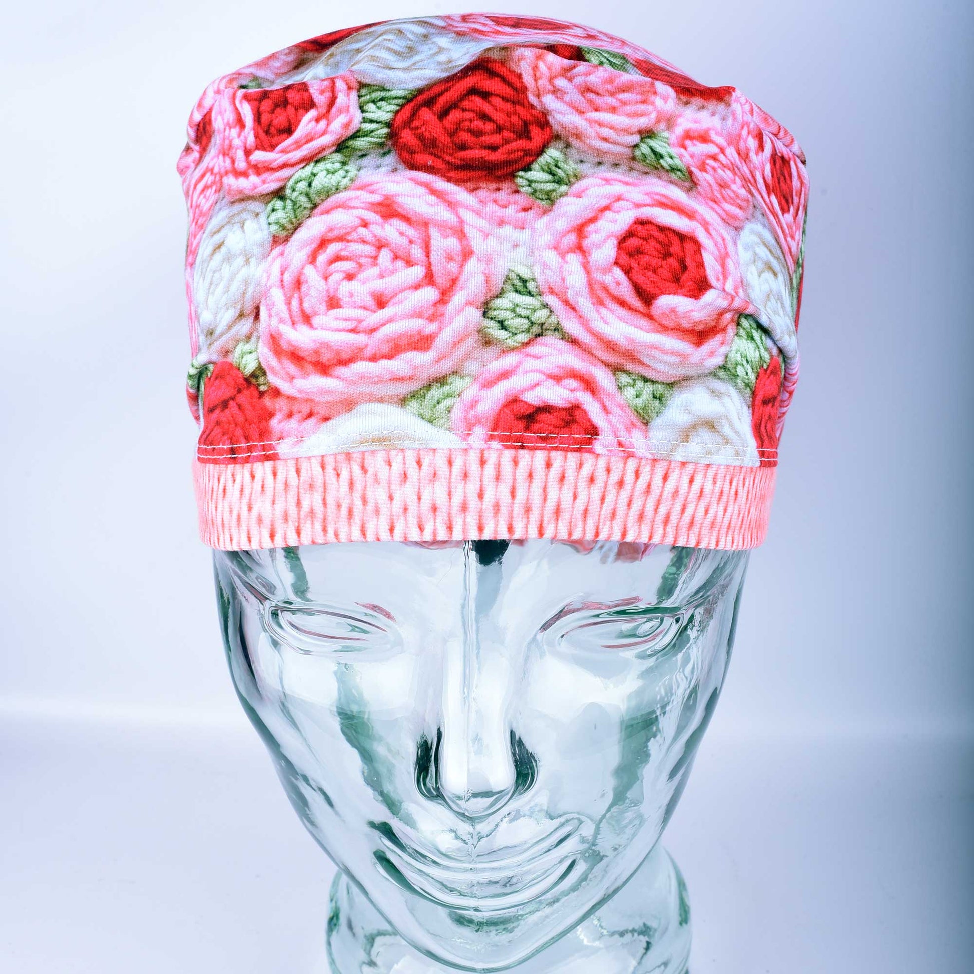 A full-coverage Euro-style scrub cap featuring a vibrant "Crochet Roses" pattern with pink and red floral motifs on a white background, displayed on a glass headform.