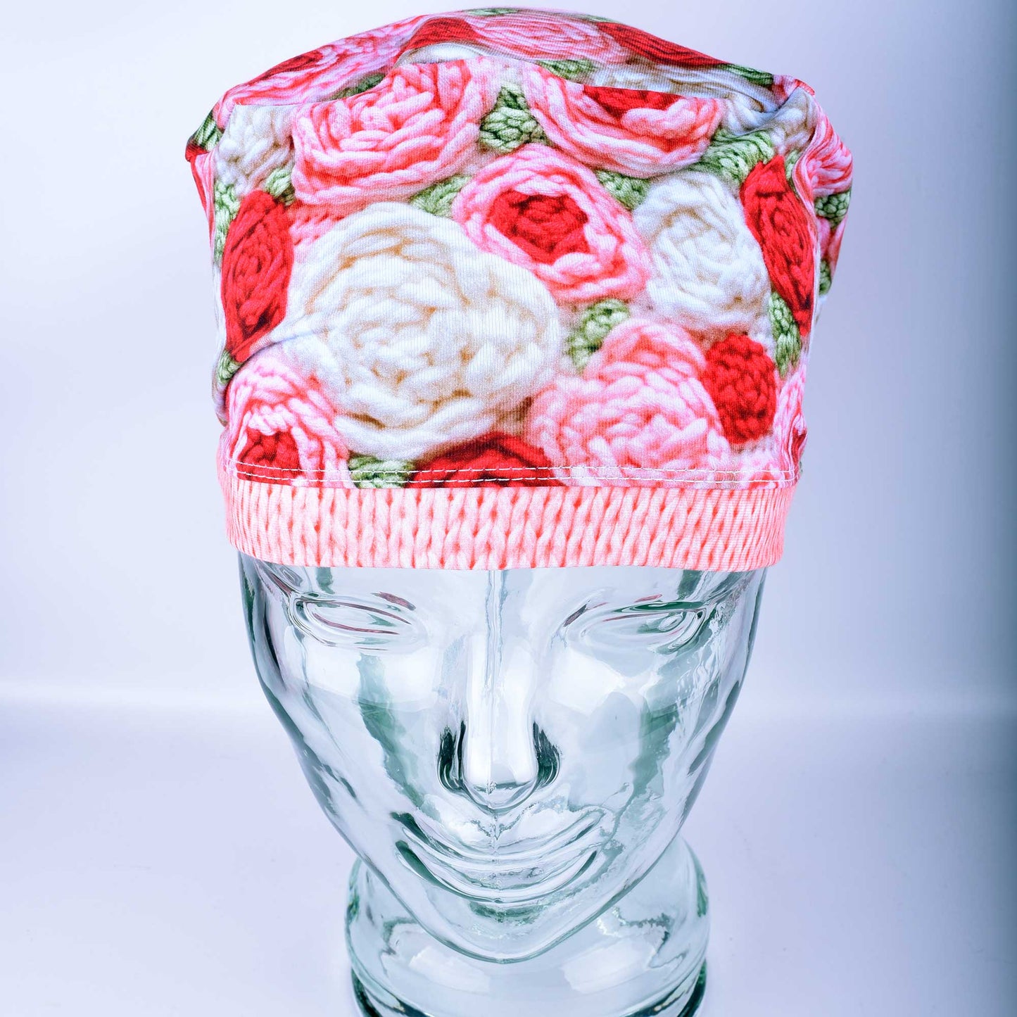 A full-coverage Euro-style scrub cap featuring a vibrant "Crochet Roses" pattern with pink and red floral motifs on a white background, displayed on a glass headform.