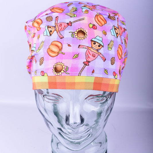 Front-facing view of a Full Coverage Scrub Cap on a clear mannequin head, showcasing a festive fall pattern with friendly cartoon scarecrows, pumpkins, and autumn leaves.