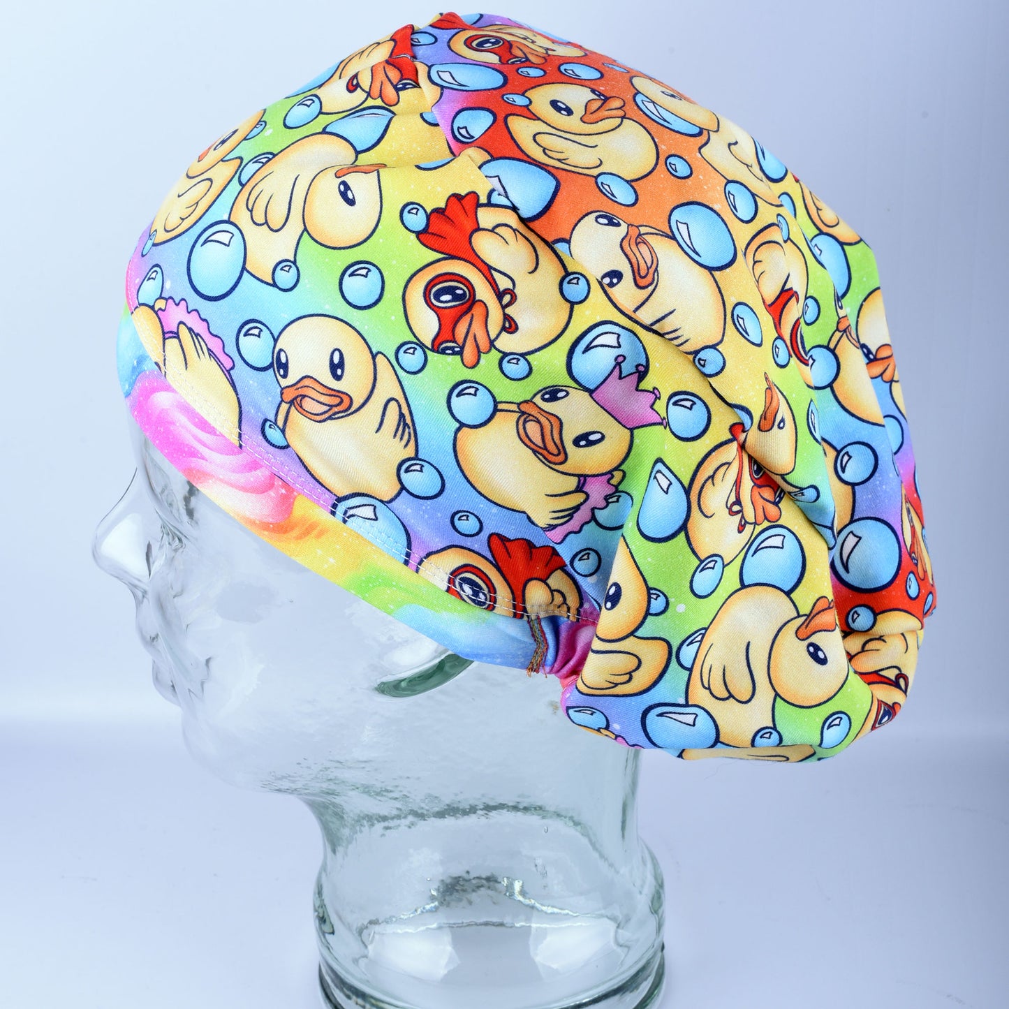 A full coverage scrub cap featuring a vibrant custom pattern of yellow rubber ducks and blue bubbles on a white background, displayed on a clear mannequin head.
