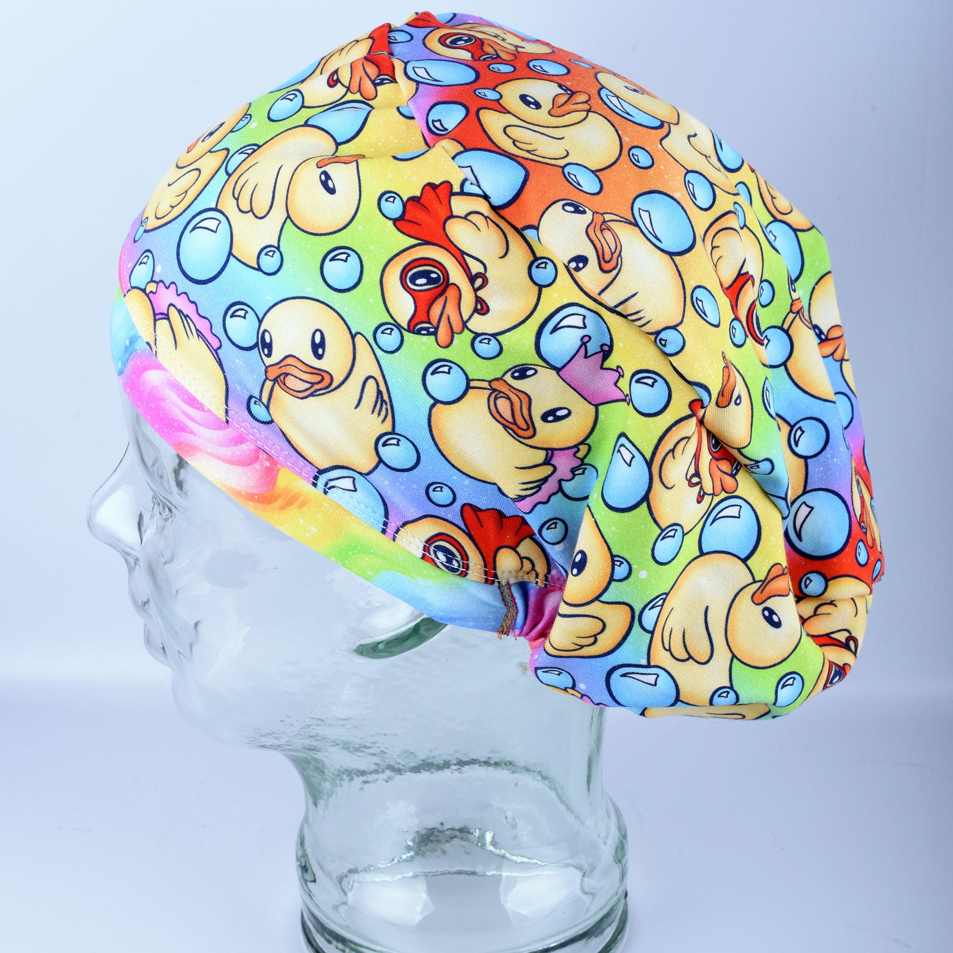 A full coverage scrub cap featuring a vibrant custom pattern of yellow rubber ducks and blue bubbles on a white background, displayed on a clear mannequin head.
