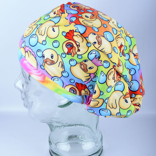 A full coverage scrub cap featuring a vibrant custom pattern of yellow rubber ducks and blue bubbles on a white background, displayed on a clear mannequin head.