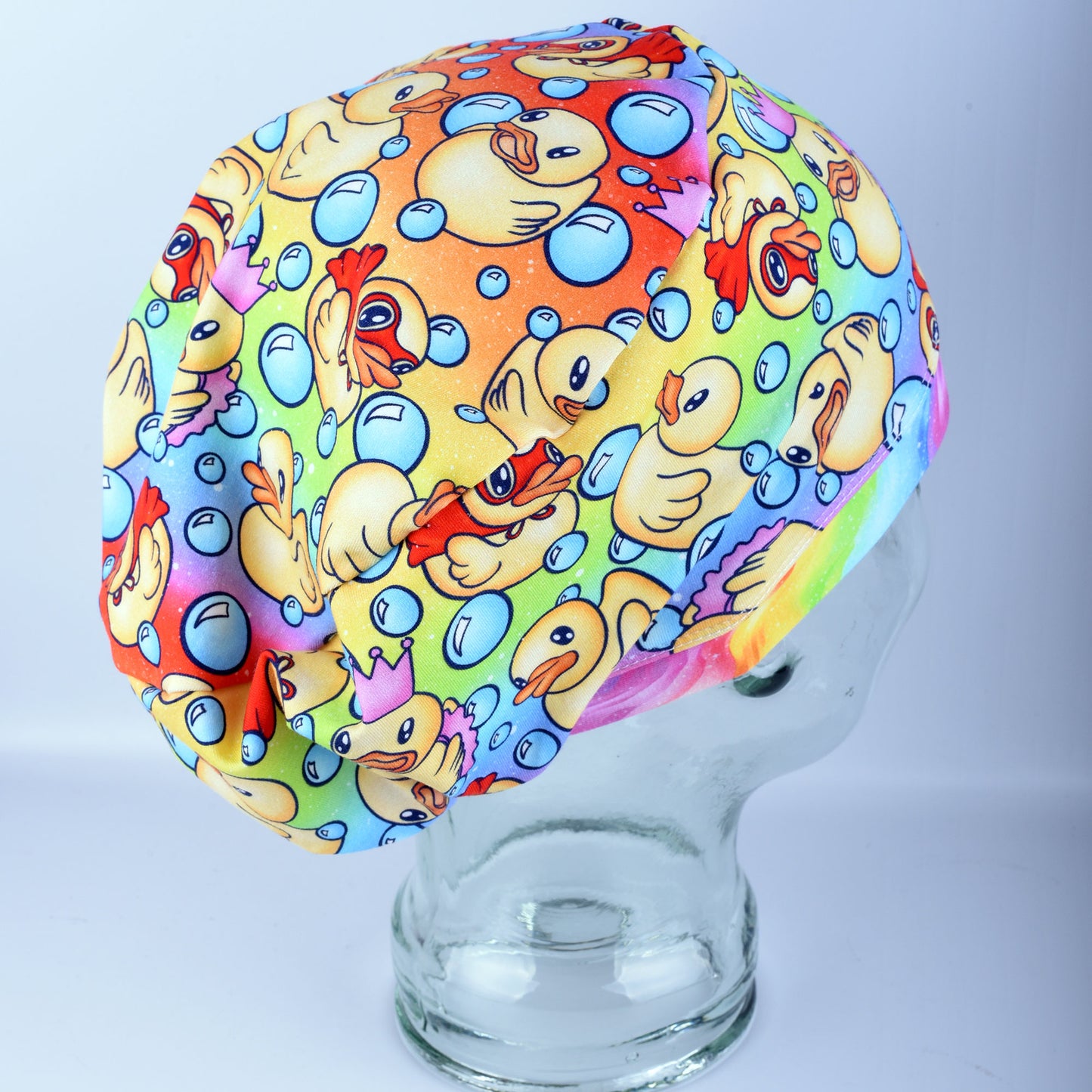 A full coverage scrub cap featuring a vibrant custom pattern of yellow rubber ducks and blue bubbles on a white background, displayed on a clear mannequin head.