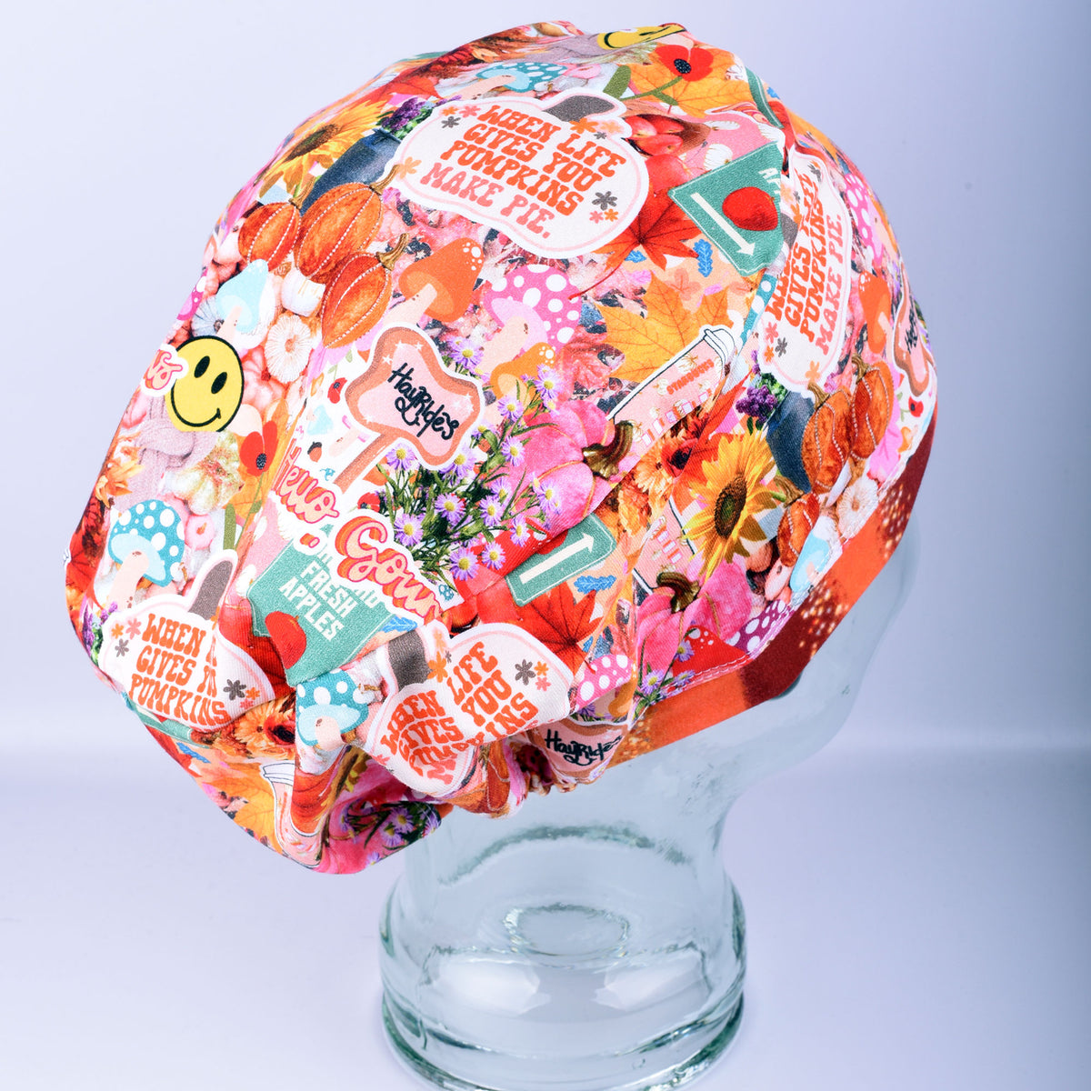 A multicolor scrub cap with a busy collage print featuring flowers, smiley faces, and text like 'WHEN LIFE GIVES YOU PUMPKINS, MAKE PIE"  displayed on a clear glass mannequin head against a light gray background.