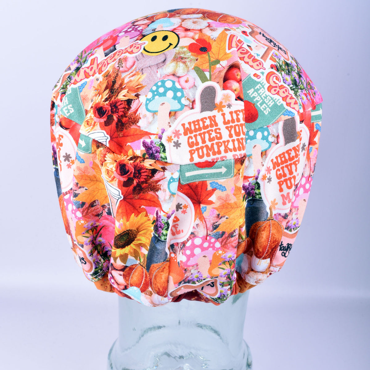 A multicolor scrub cap with a busy collage print featuring autumn leaves, pumpkins, flowers, and text like 'When life gives you pumpkins make pie', displayed on a clear stand against a light gray background.