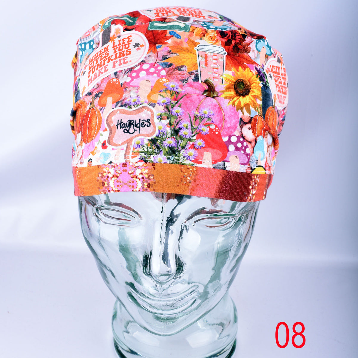 A colorful patterned surgical scrub cap displayed on a clear glass mannequin head against a light gray background. The cap features a busy collage print with autumn leaves, pumpkins, flowers, and text elements, finished with a glittery orange-red band.