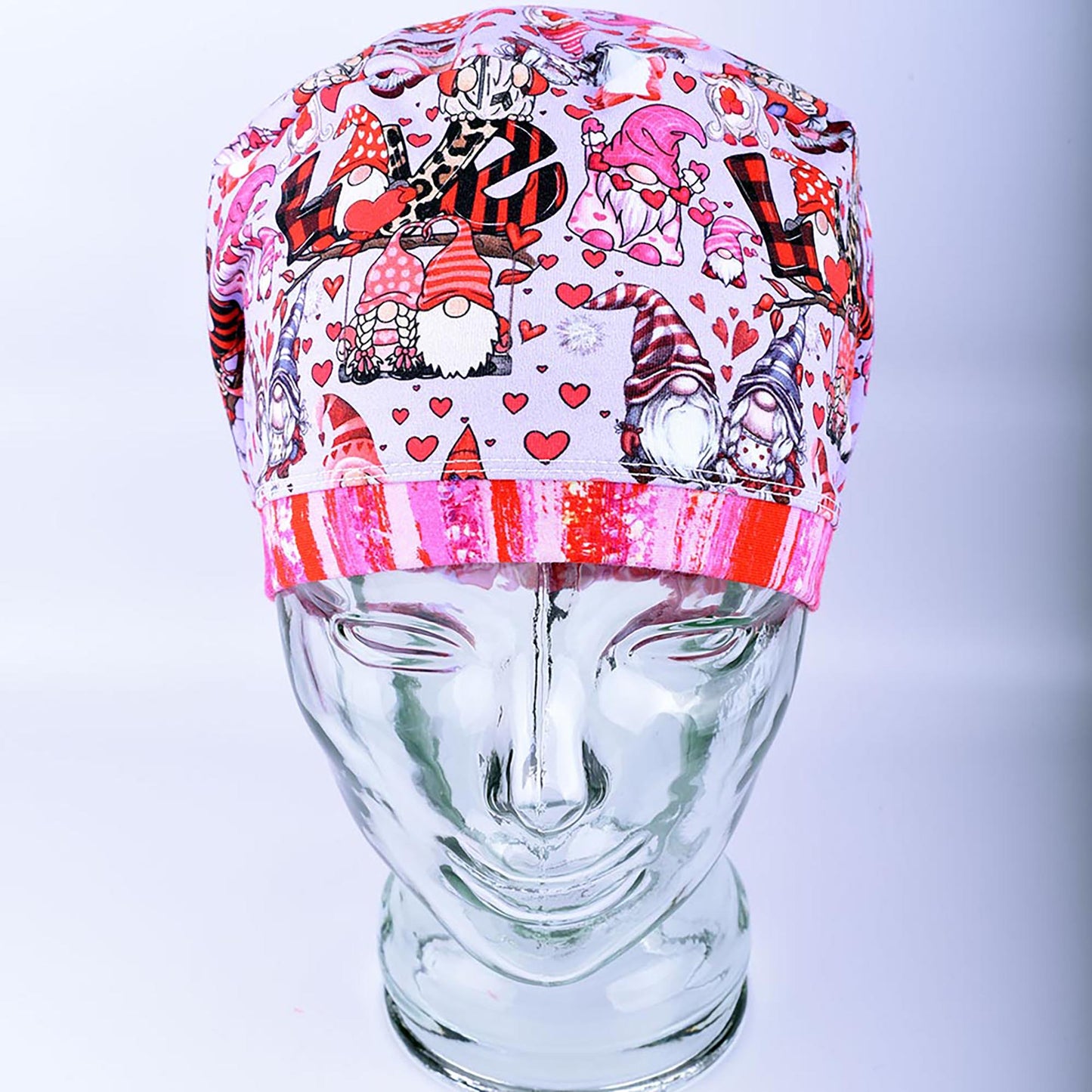 A full coverage Euro-style scrub cap featuring a whimsical Gnome Love print with pink and red hearts, folded neatly on a clear glass mannequin stand.
