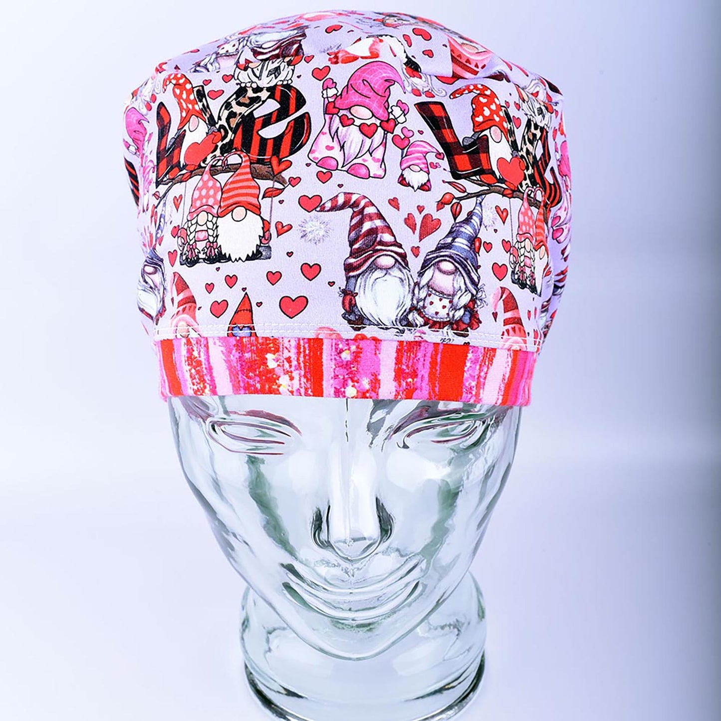 A full coverage Euro-style scrub cap featuring a whimsical Gnome Love print with pink and red hearts, folded neatly on a clear glass mannequin stand.