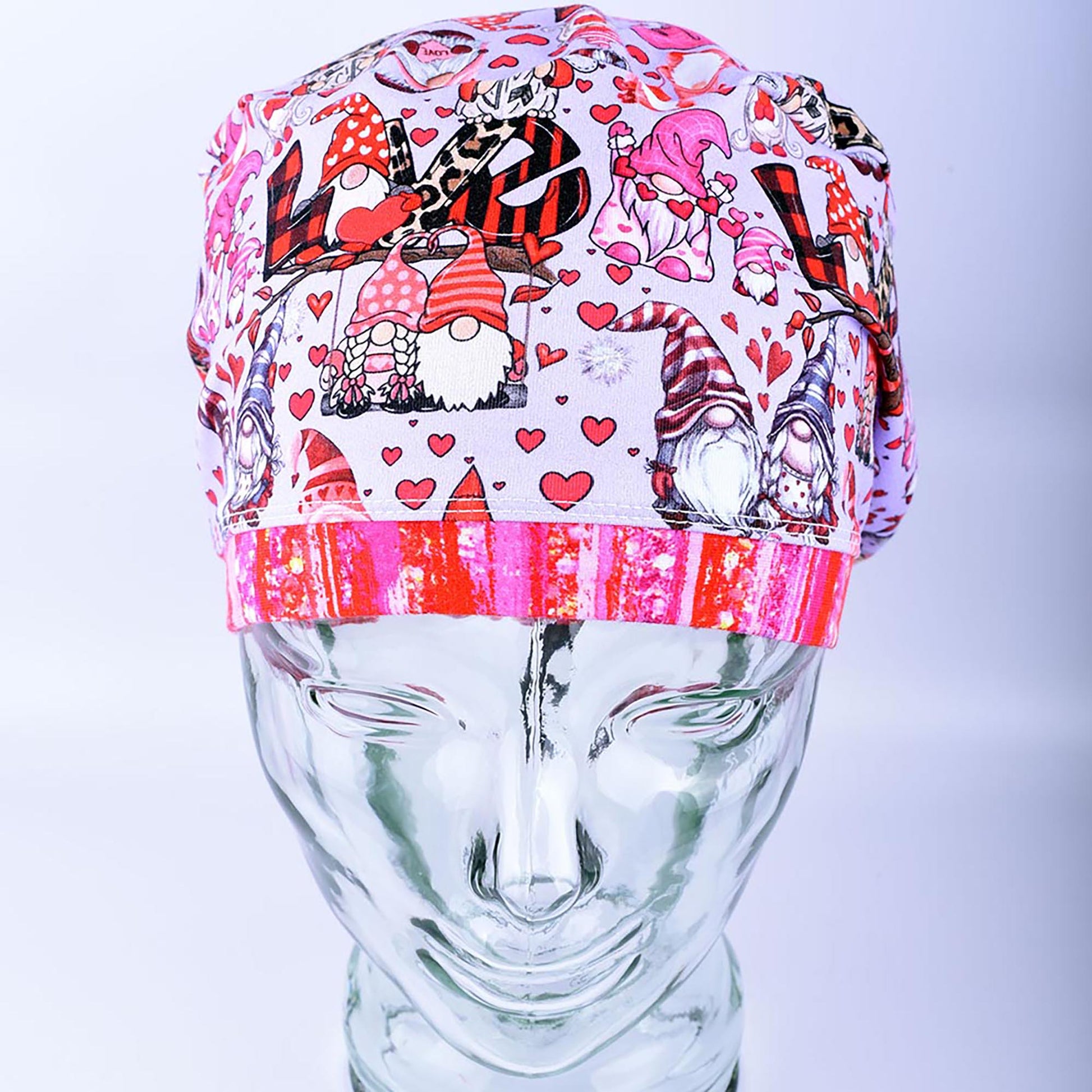 A full coverage Euro-style scrub cap featuring a whimsical Gnome Love print with pink and red hearts, folded neatly on a clear glass mannequin stand.