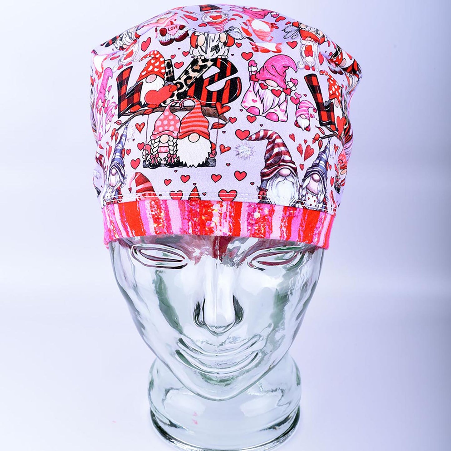 A full coverage Euro-style scrub cap featuring a whimsical Gnome Love print with pink and red hearts, folded neatly on a clear glass mannequin stand.