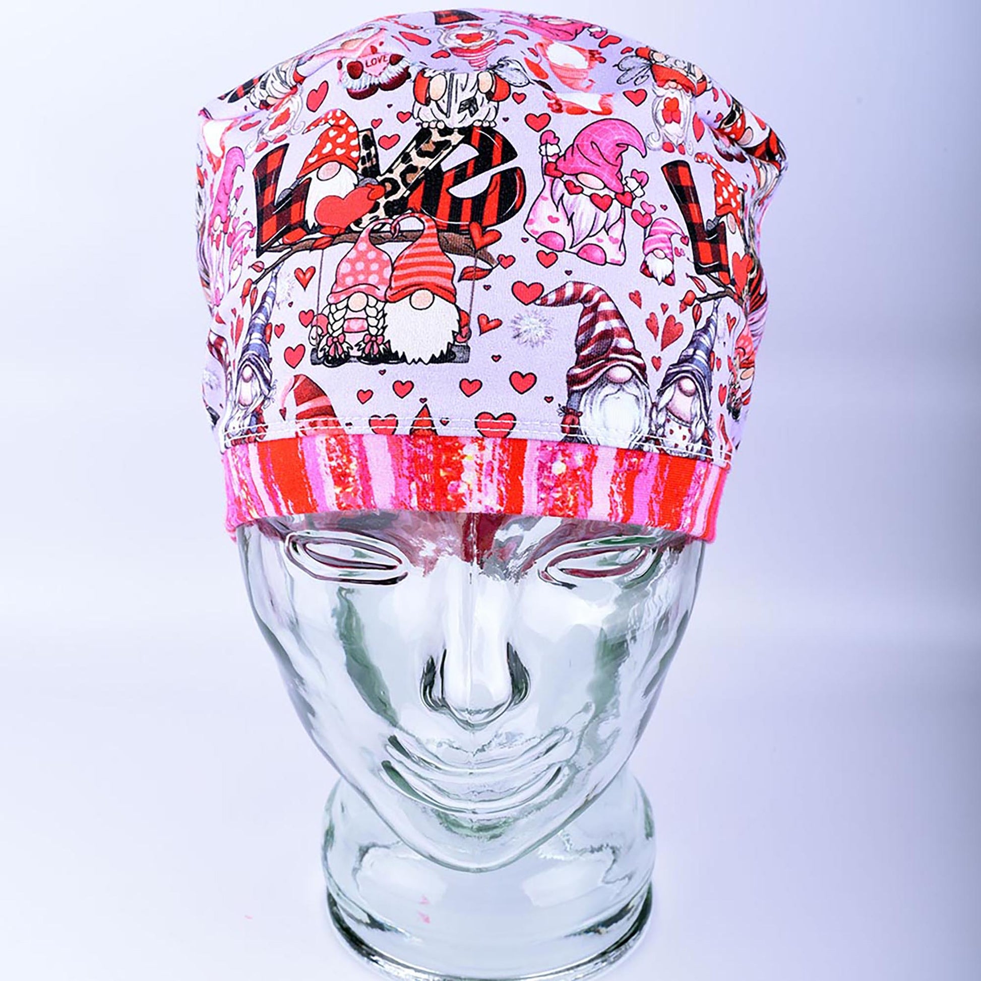 A full coverage Euro-style scrub cap featuring a whimsical Gnome Love print with pink and red hearts, folded neatly on a clear glass mannequin stand.