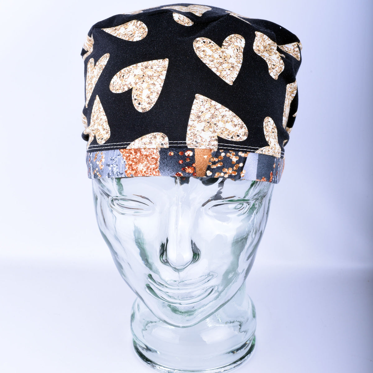 A black fabric scrub cap with a pattern of glittery gold hearts and a patterned elastic band, displayed on a clear glass mannequin head against a light gray background.