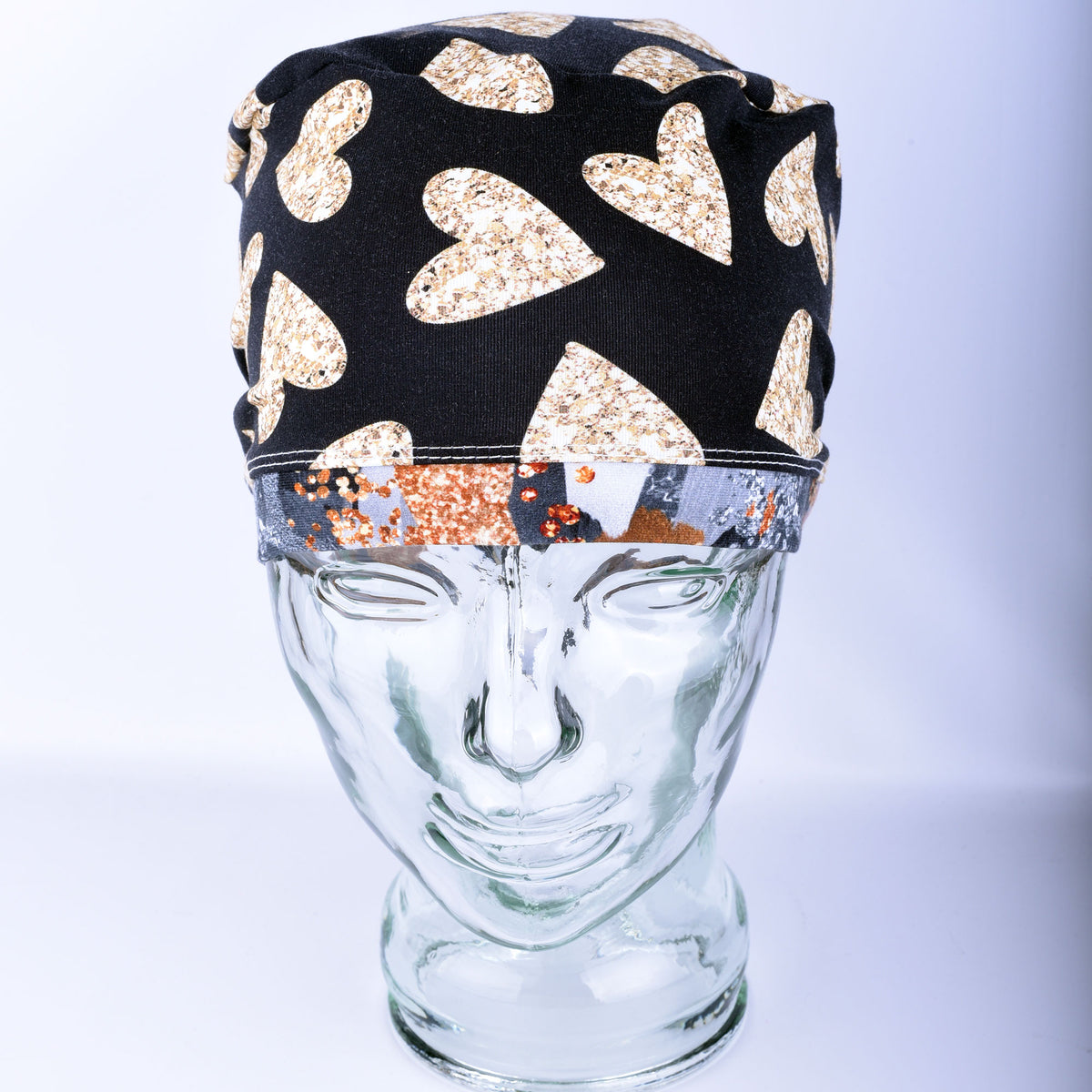 A black fabric scrub hat with a pattern of glittery gold hearts, displayed on a clear glass mannequin head against a light gray background.