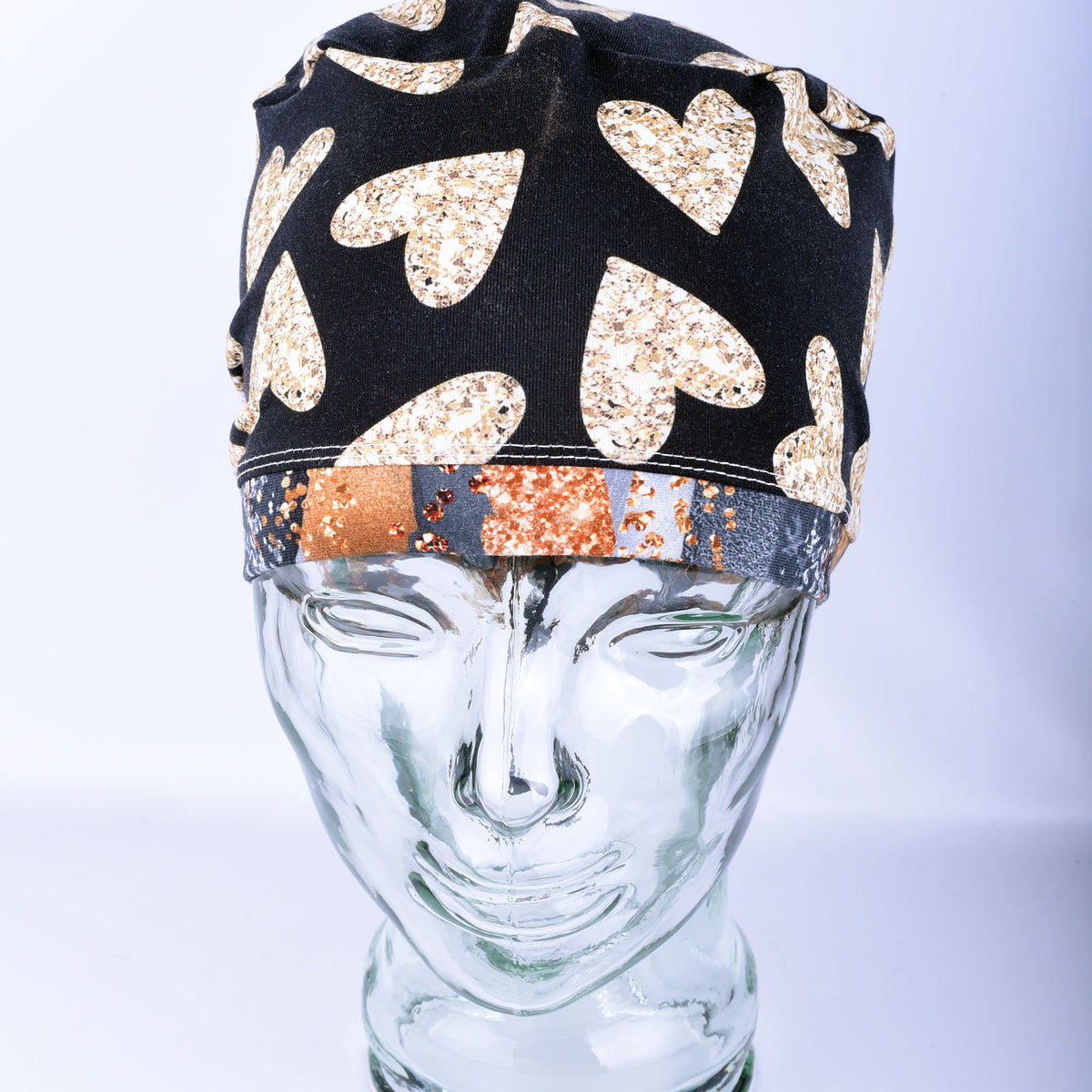 A black fabric surgical-style cap with large glittery gold hearts and a patterned band, displayed on a clear glass mannequin head against a light gray background.