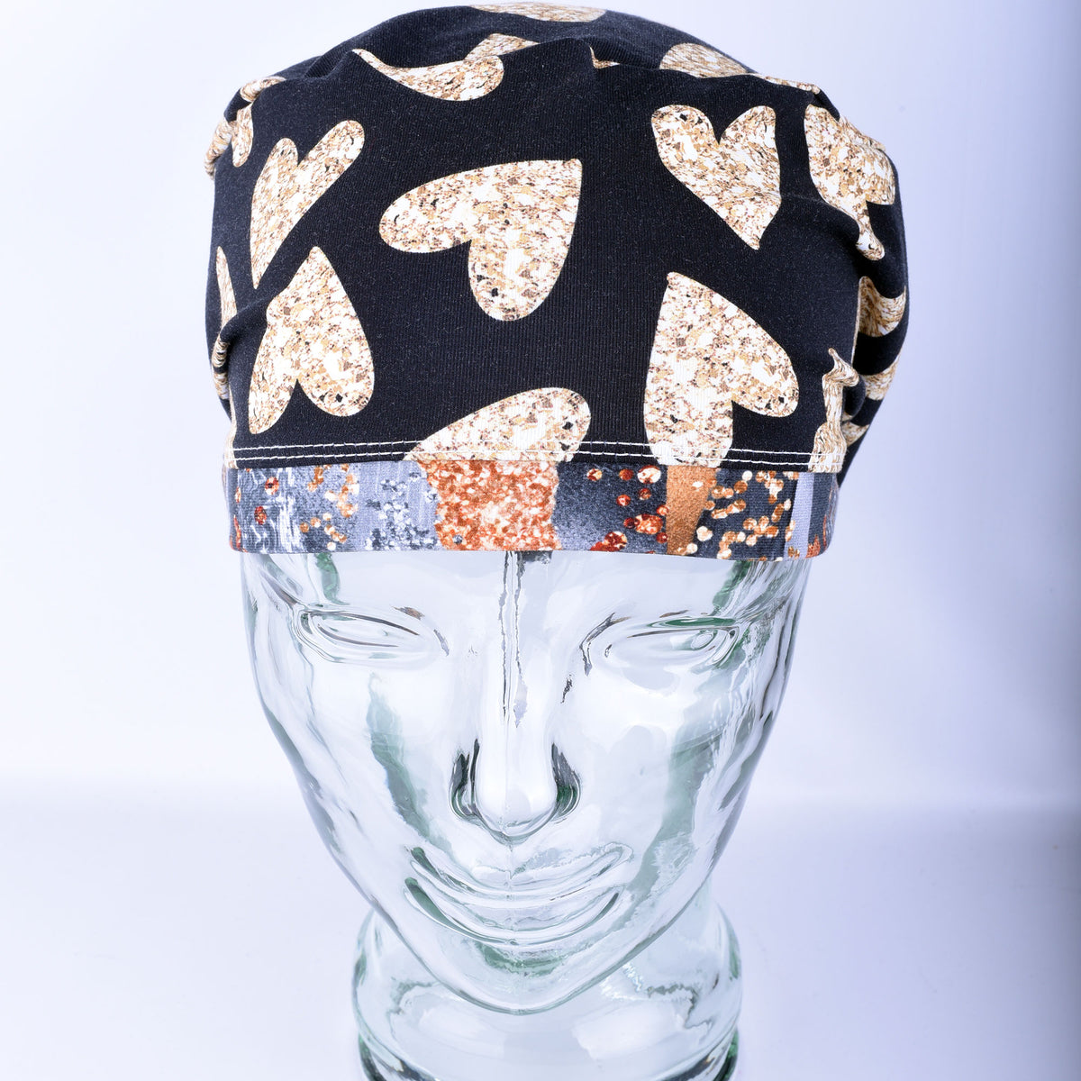 A black fabric surgical cap with a pattern of glittery gold hearts and a multicolor patterned band, displayed on a clear glass mannequin head against a light gray background.