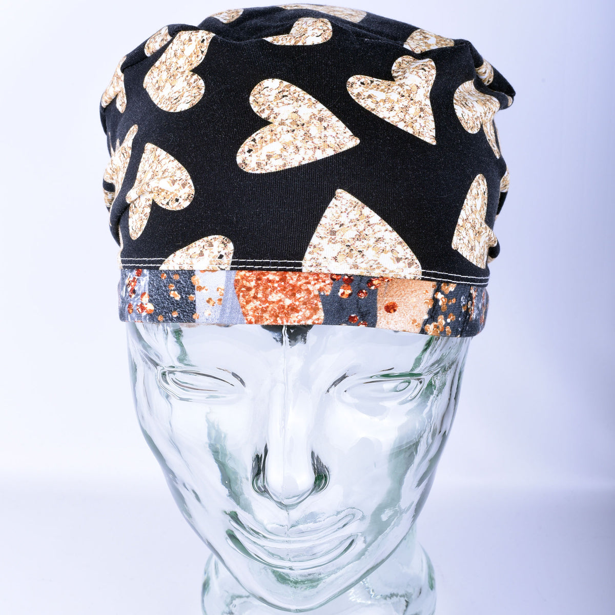 A black fabric head covering or cap with large glittery gold hearts and a multicolor patterned band, displayed on a clear glass mannequin head against a light gray background.