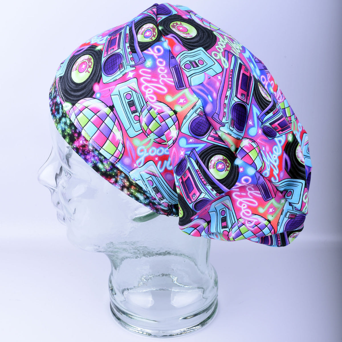 A colorful fabric scrub cap with a retro 1980s disco pattern, featuring boomboxes, turntables, and neon lights, displayed on a clear glass mannequin head against a light gray background.