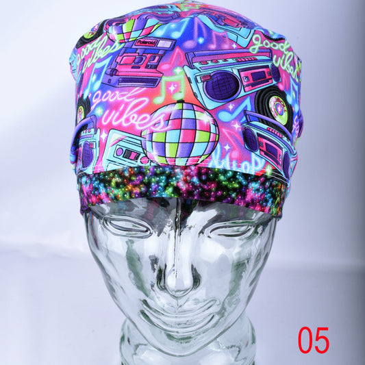 A neon 80s disco-themed fabric head covering or cap displayed on a clear glass mannequin head. The cap features a vibrant multicolor pattern with boomboxes, a disco ball, cassette tapes, and 'good vibes' text.