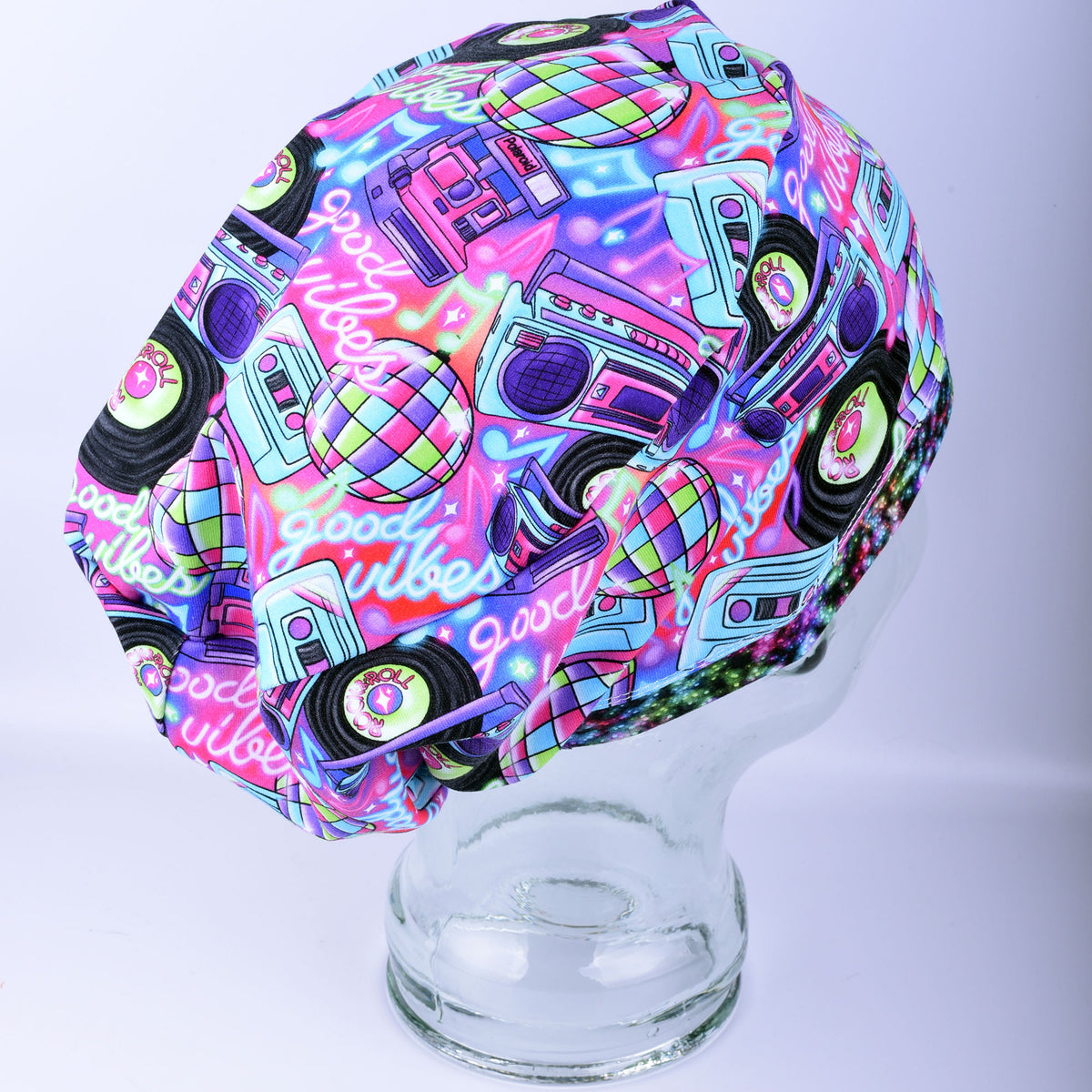 A neon pink and blue scrub cap with a retro 80s disco-themed print, featuring boomboxes, cassettes, and a disco ball, displayed on a clear glass mannequin head against a light gray background.