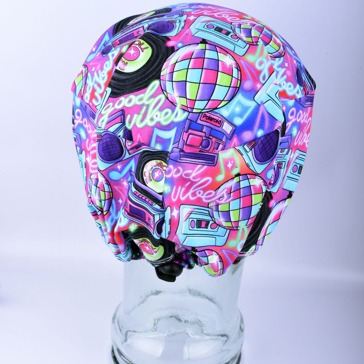 A vibrant, multicolor fabric headband with a retro 1980s/90s 'Good Vibes' theme, featuring neon graphics, cassette tapes, boomboxes, and disco balls, displayed on a clear glass stand.