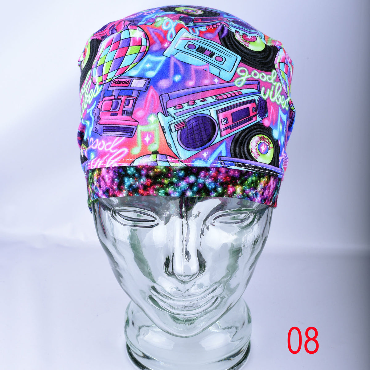 A neon multicolor medical cap with a retro boombox, cassette tape, and cassette vinyl graphic print, displayed on a clear glass mannequin head. The headband features a glittery multicolor elastic band at the bottom.