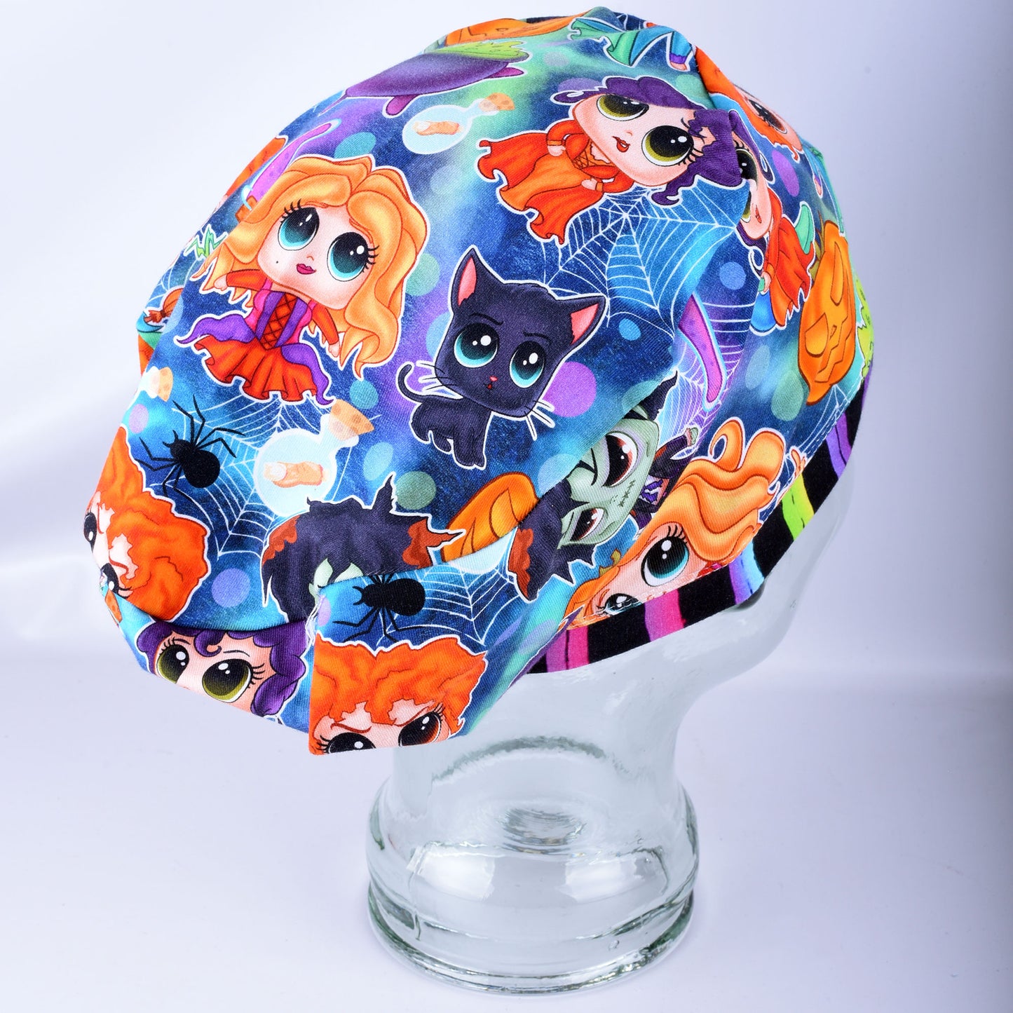 A multicolor Halloween-themed scrub cap featuring cartoon characters like a black cat, orange-haired dolls, and spiders on a blue spiderweb background, displayed on a clear glass mannequin head.