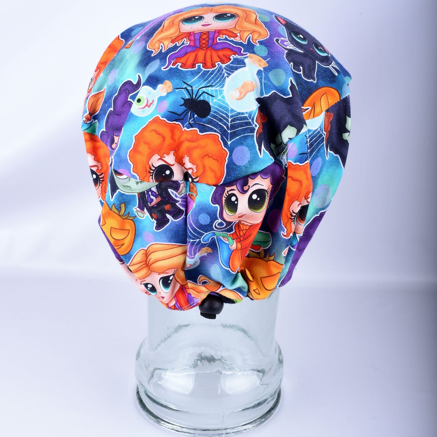 A Halloween-themed medical scrub cap with a colorful cartoon print, displayed on a clear glass mannequin head against a white studio background.