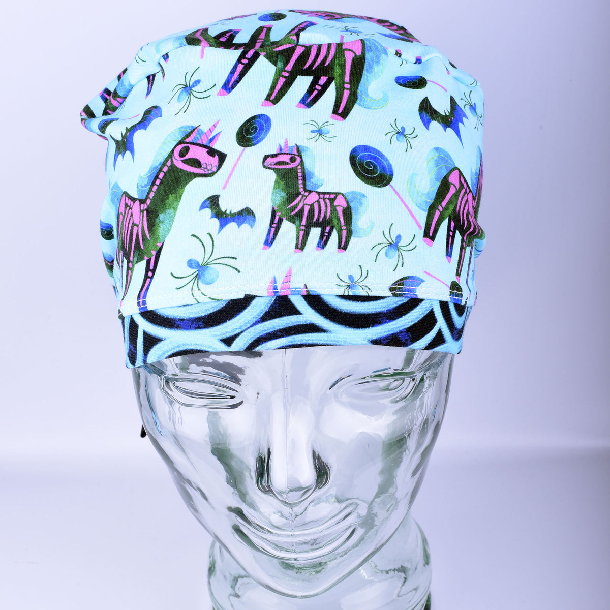 Front view of unicorn skeleton scrub cap on a clear mannequin head showing full coverage fit and light blue background pattern.