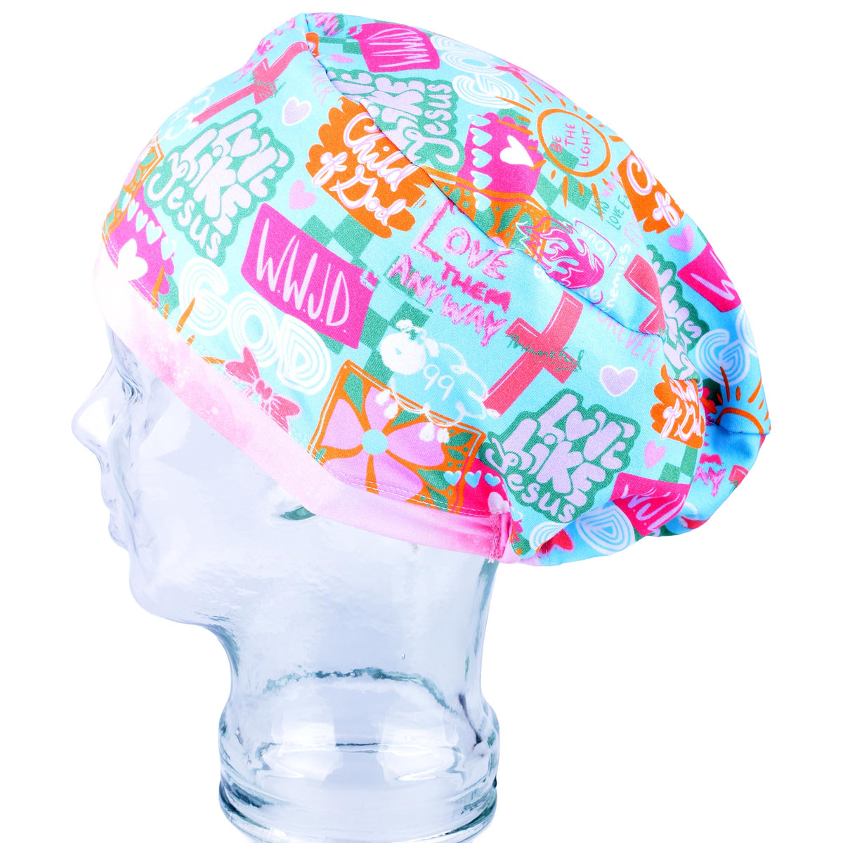 A multicolor scrubs cap with a busy graphic pattern featuring crosses, hearts, and religious text, displayed on a clear glass mannequin head against a white background.