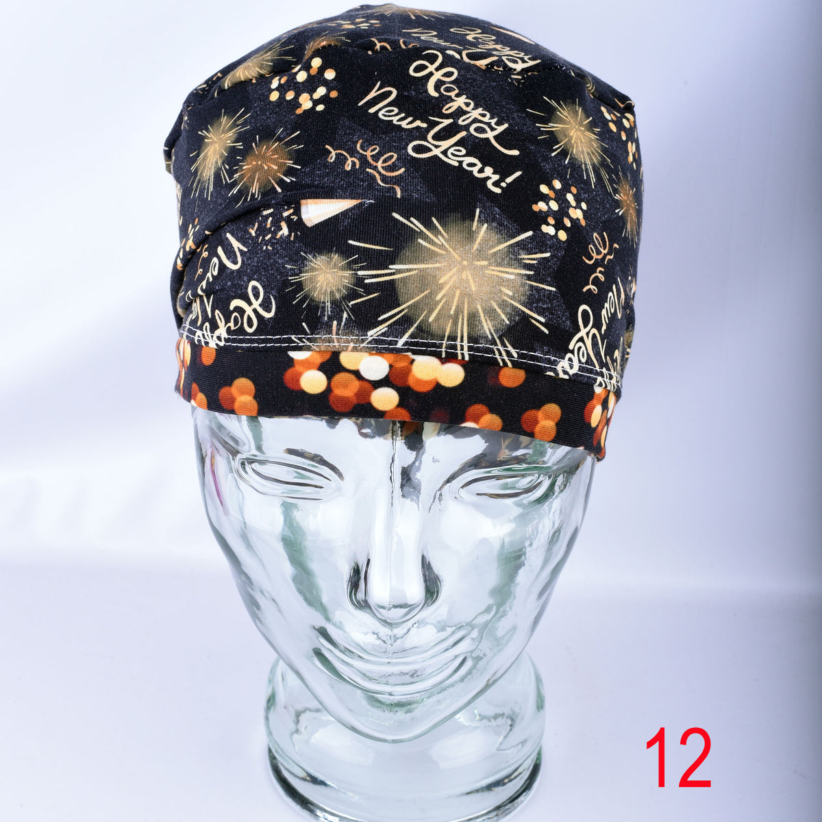A black fabric head covering or cap with a New Year's Eve theme, featuring gold fireworks, 'Happy New Year!' script, and orange/gold confetti, displayed on a clear glass mannequin head. A red number '12' is visible in the bottom right corner.